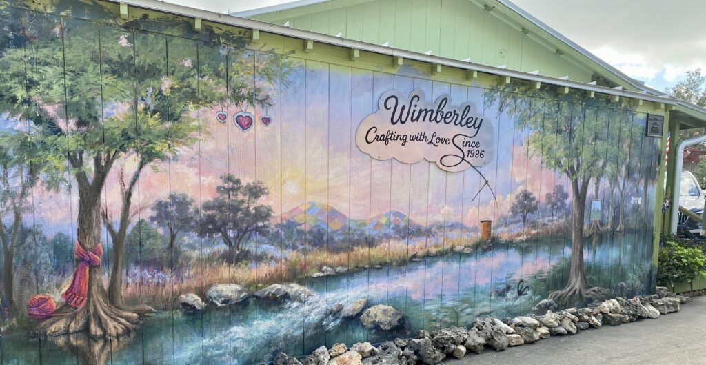 Outdoor mural on a building exterior reading "Wimberley – Crafting with Love Since 1986," depicting a scenic landscape with trees, a creek, yarn, and quilts
