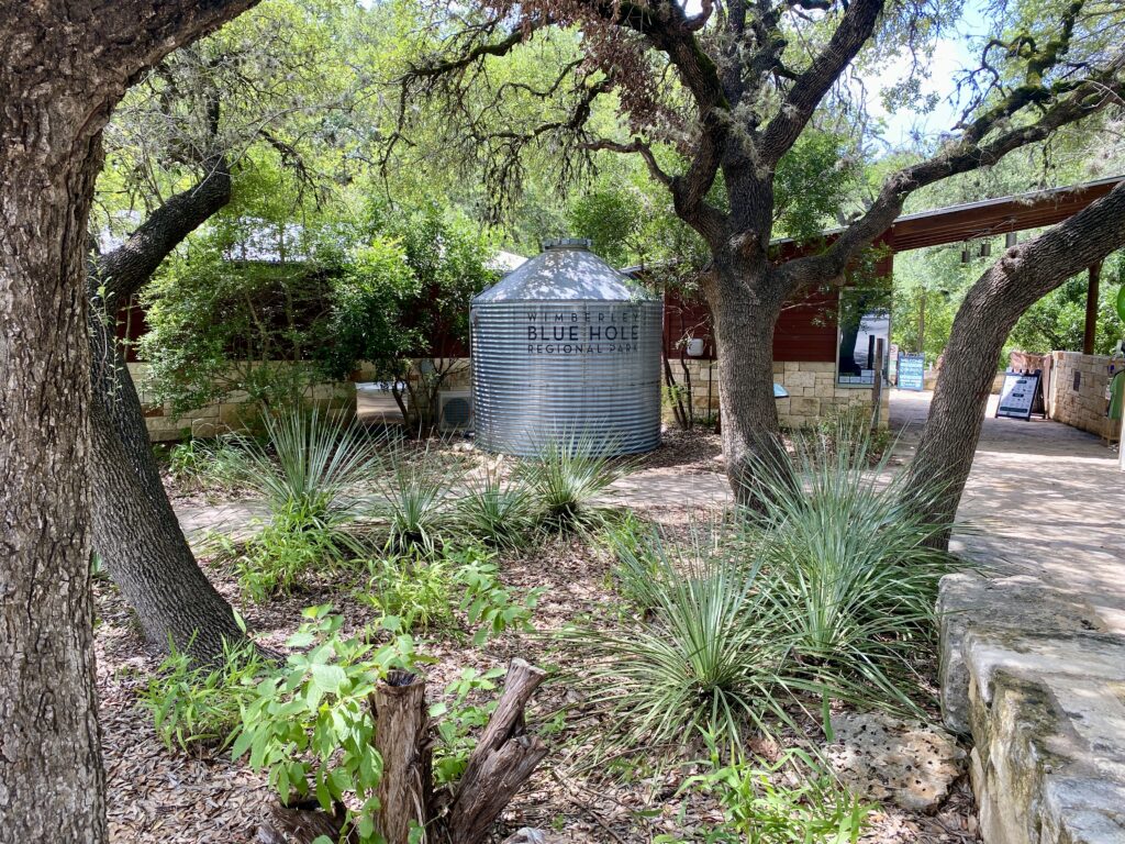 Entrance area of Wimberley Blue Hole Regional Park, with a metal tank bearing the park name surrounded by native plants and oak trees