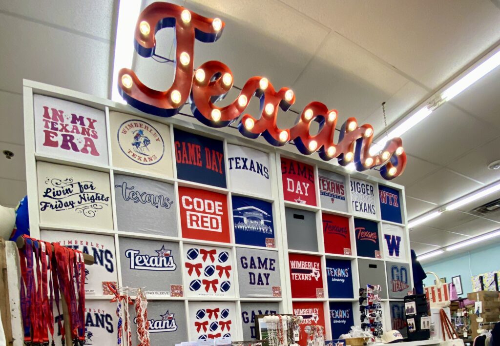 Wimberley Texans spirit wear display wall with a lit marquee Texans sign above shelves of red, white, and blue graphic tees at Ace Hardware and Gifts.